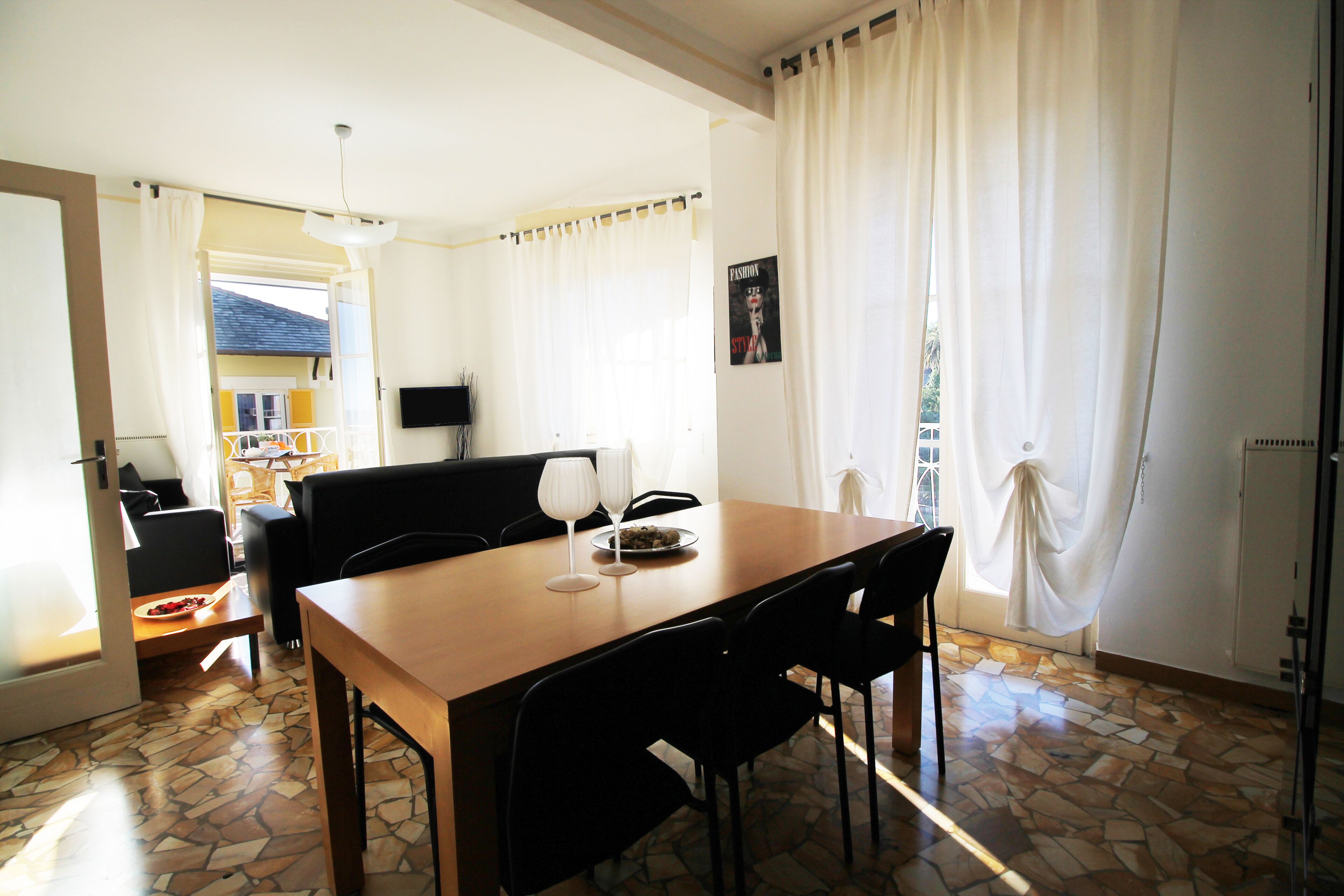 A dining area with a wooden table, black chairs, and a small table with a bowl and glasses. There is a TV on the wall and a window with curtains.