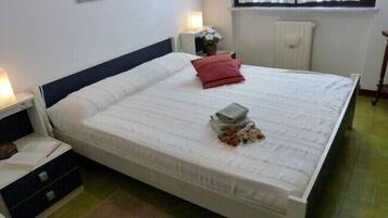 Cama bem arrumada com colchão branco e almofada vermelha, mesa de cabeceira com abajur e janela com cortinas.