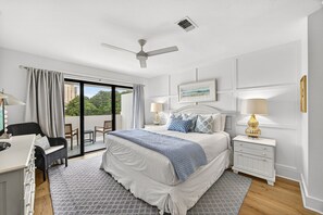 A bedroom with a bed, two bedside tables with lamps, a ceiling fan, a window with curtains, and a view of greenery outside.