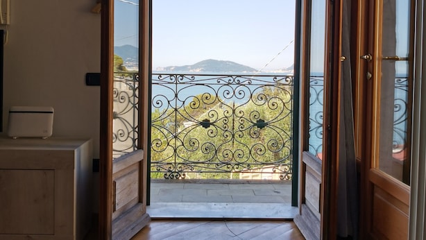 An open door leading to a balcony with a metal railing, wooden floor, and a view of a mountain and sea.