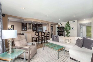 Ein modernes Wohnzimmer mit einer Couch, Sesseln, einem Glastisch und einem Essbereich mit Stühlen sowie einer Küche im Hintergrund.