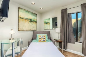 Habitación de hotel con cama, mesita de vidrio, lámpara, cuadro, espejo y ventana con cortinas.