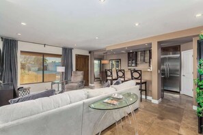 Una sala de estar moderna con sofá, mesa de centro de vidrio, comedor con sillas, cocina con refrigerador y un ventanal con persianas.