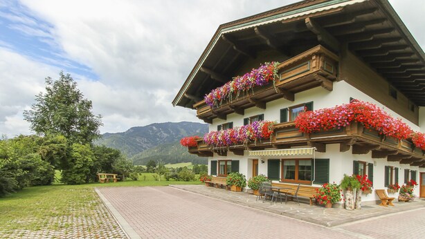 A traditional chalet with flower boxes on the balconies, a paved driveway, and a bench outside. - Schneiderbauer (Waidring)