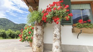 Two large wooden planters with red flowers on a stone-paved porch, a window with green shutters, and a mountainous backdrop. - Schneiderbauer (Waidring)