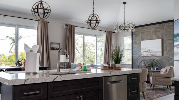 A modern kitchen with dark cabinetry, a marble countertop, and stainless steel appliances. There are two pendant lights, a large window with curtains, and a view of greenery outside. - New 4BR Oasis: Walk to Beach, Pool, Pet-Friendly! (Fort Myers Beach)
