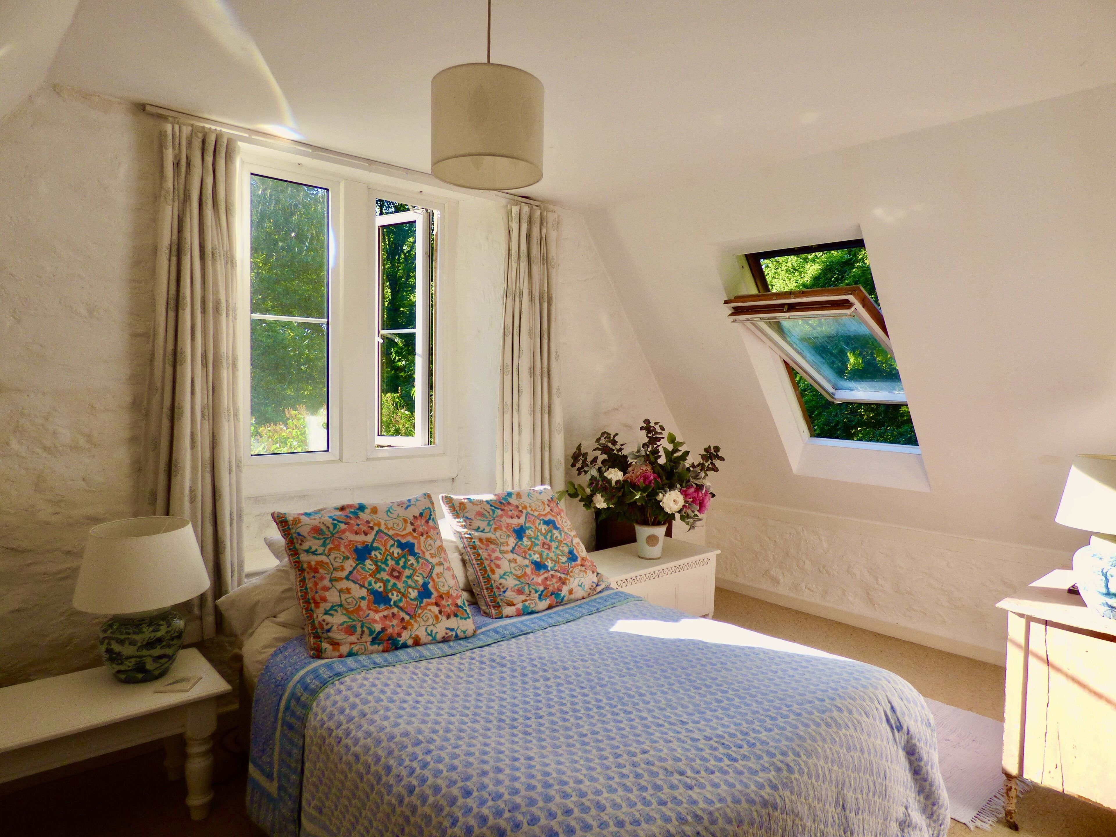 Une chambre à coucher comprenant un lit, une table de chevet, une lampe, une fenêtre avec des rideaux, un vase à fleurs et une lucarne.