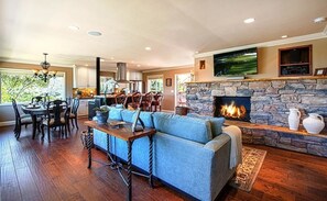 A spacious living room with a stone fireplace, a large TV mounted above it, and a comfortable seating area with a sofa and chairs. - Monarch - Pacific Grove Triplex - Ocean View! (Pacific Grove)