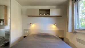 A bedroom with a bed, bedside tables, a built-in shelf with decorative items, a window with curtains, and a ceiling light. - Rent mobile home 6pers 40 m2 from 250 € per week (pont aven)
