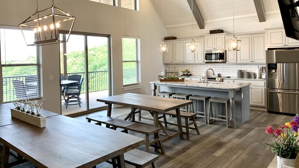 A spacious kitchen with white cabinets, a stainless steel refrigerator, and a large island with a sink. There are wooden tables and benches, and a balcony with outdoor seating.