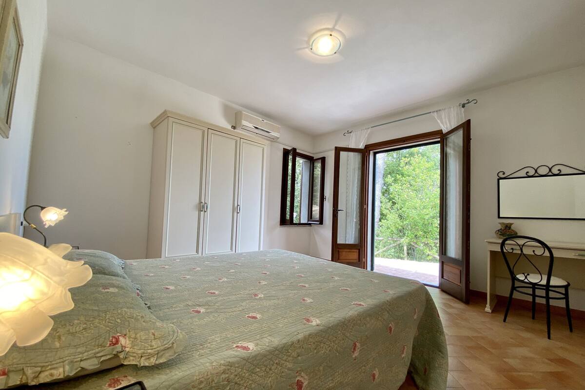 A bedroom with a bed, a desk, a wardrobe, and a window with a view of greenery.