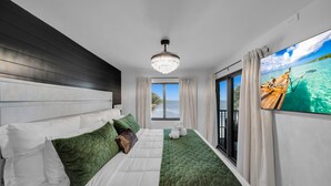 Een moderne hotelkamer met een groot bed, een kroonluchter, uitzicht op de oceaan door een schuifdeur en een televisiescherm waarop een boot op het water te zien is.