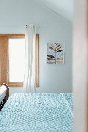 Un dormitorio con una cama, una ventana con cortinas y una obra de arte con diseño de hojas, colgada en la pared.