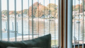 A view through a window with vertical blinds, featuring a lake with boats, docked houses, and autumn-colored trees.