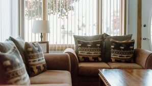 A living room with a sofa set, patterned cushions, a side table with a lamp, and a window with blinds.