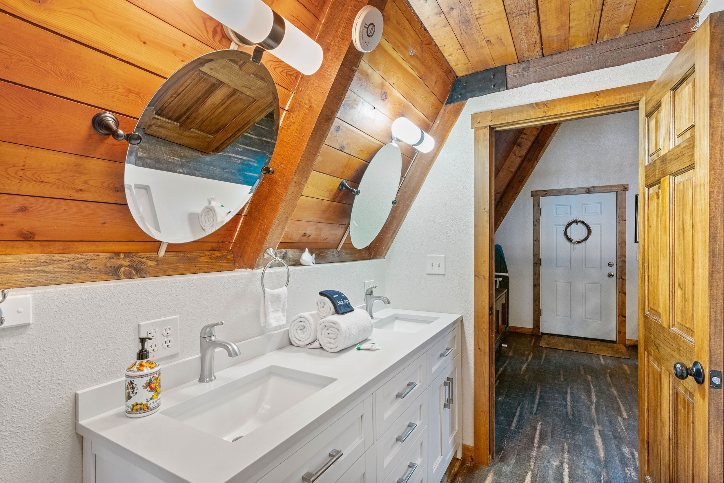 A bathroom with wooden paneling, a large mirror, two sinks, and a door leading to another room.