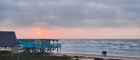 Une maison de plage avec une terrasse donnant sur l’océan, une chaise sur le sable et un oiseau dans le ciel.