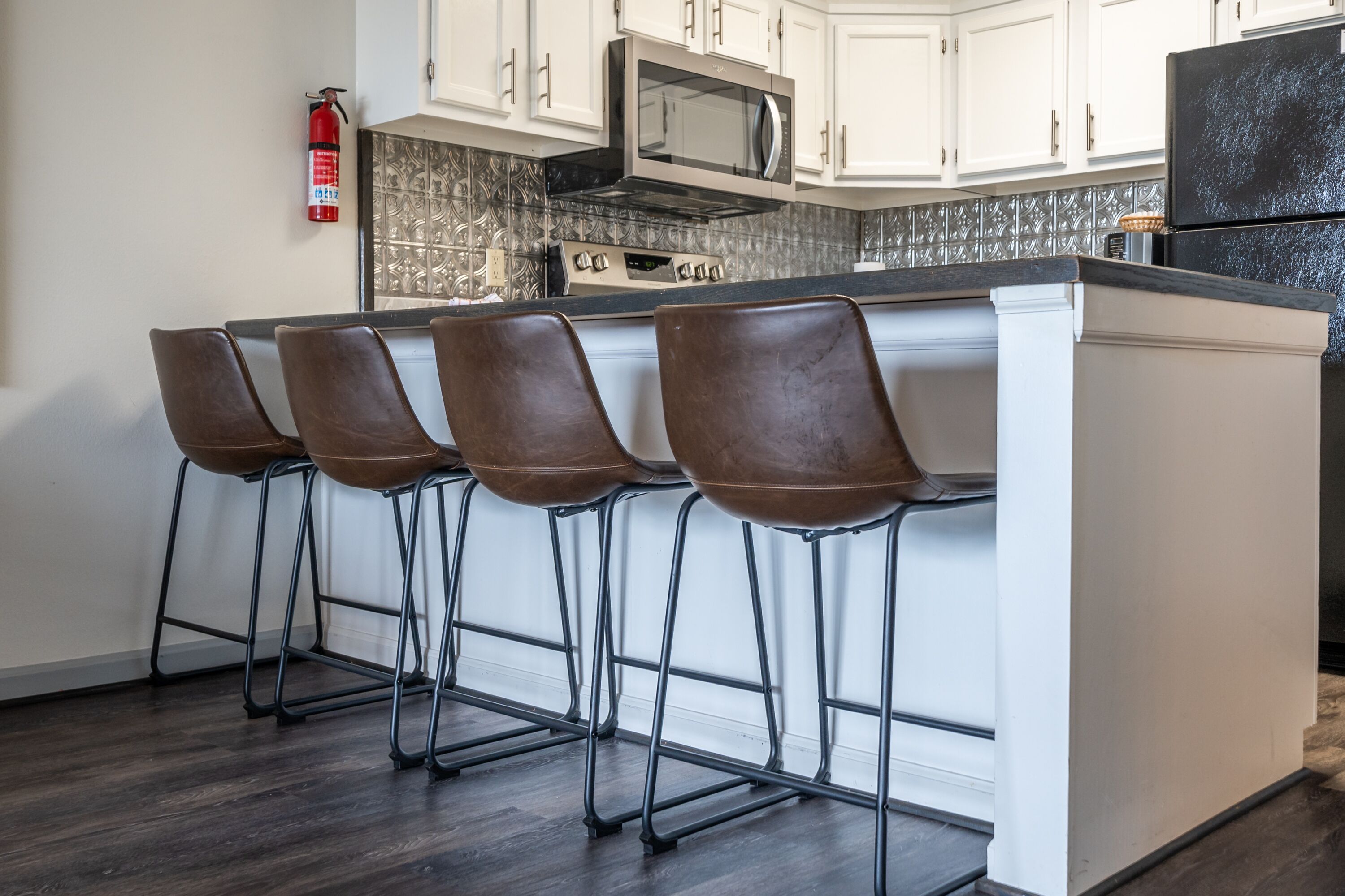 Una cocina con una isla central, cuatro taburetes de bar, un microondas, una estufa y un extintor.