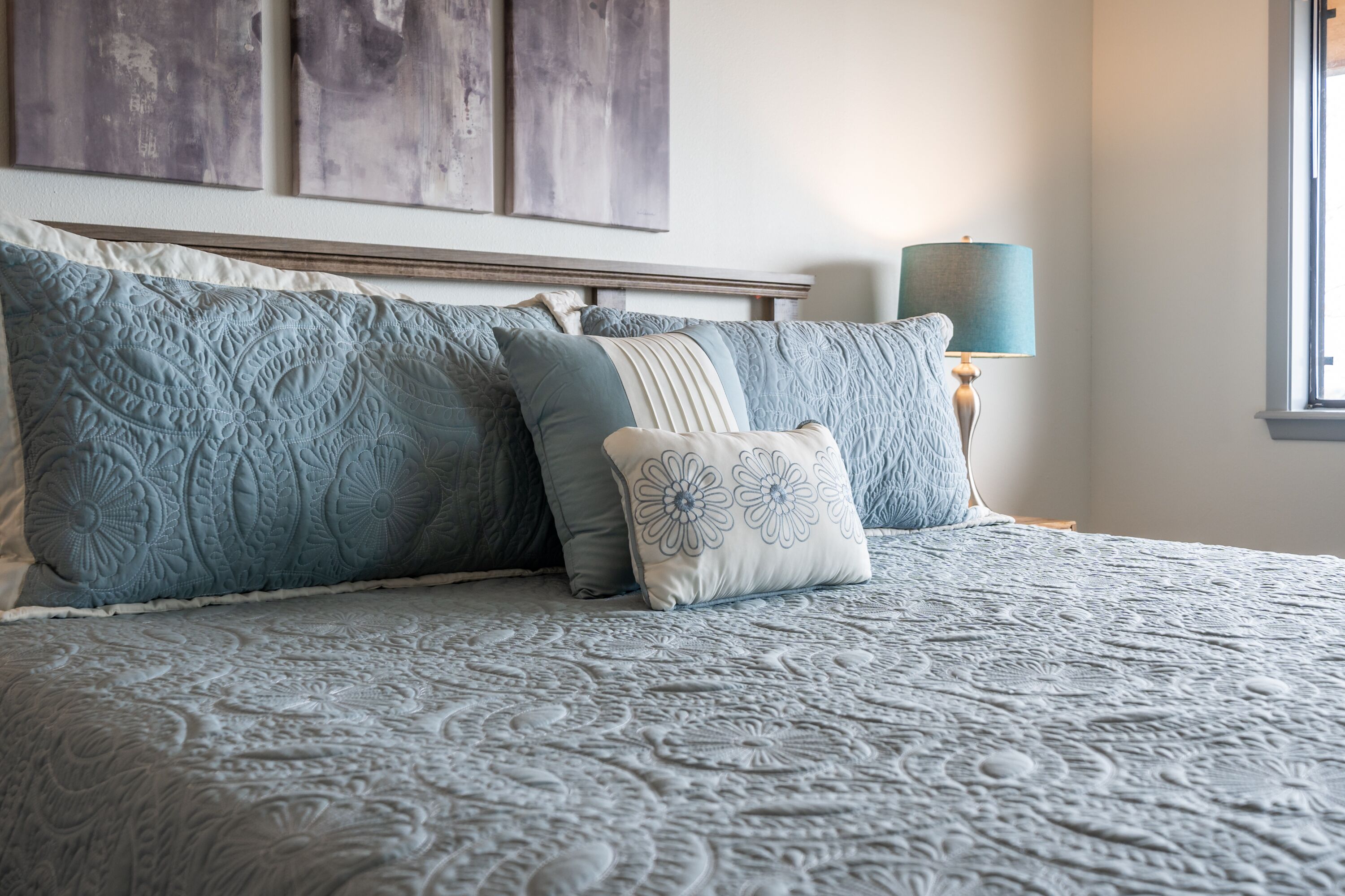 A bed with a patterned bedspread and multiple pillows, a bedside lamp, and a window with a view.