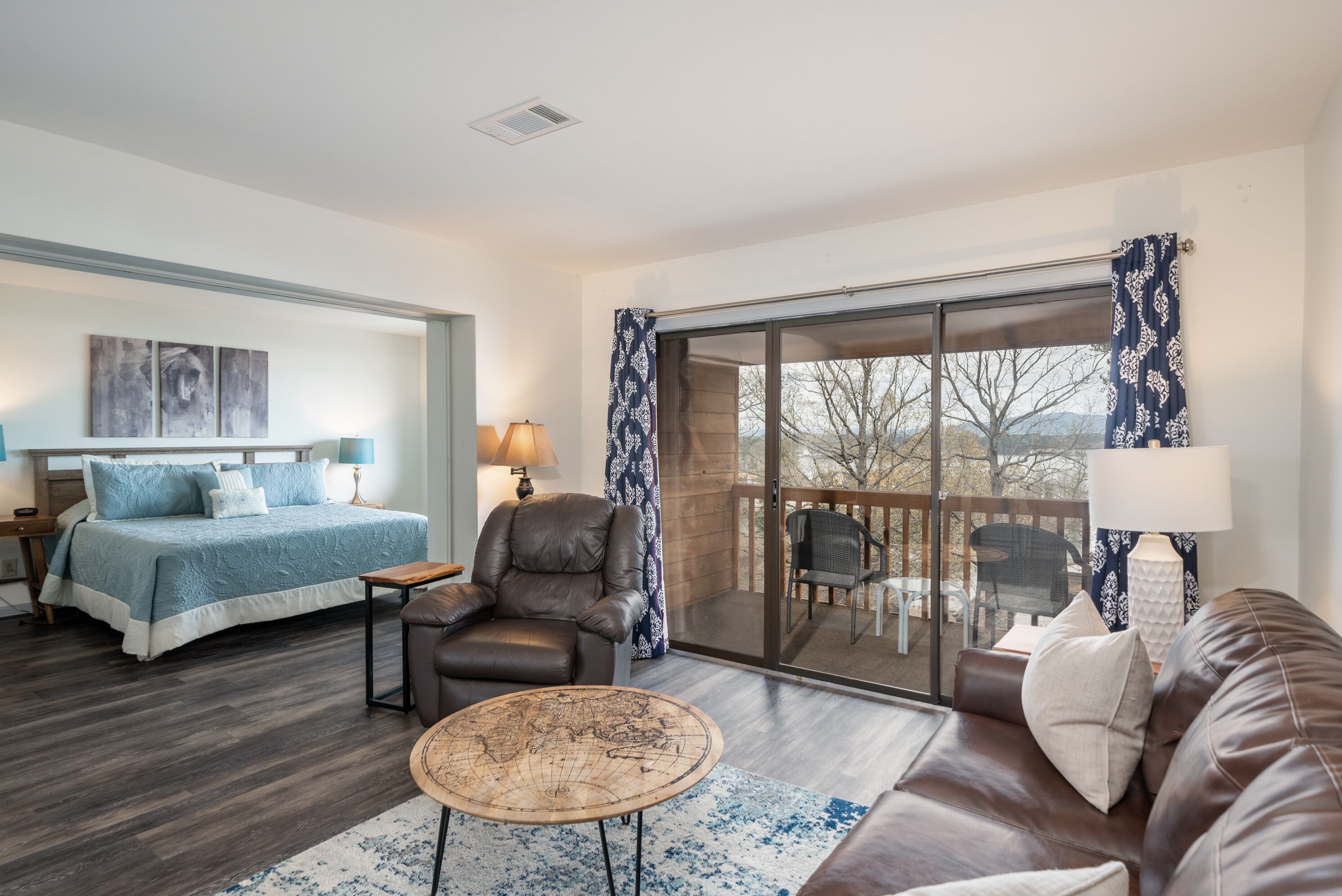 A modern hotel room with a large bed, a leather armchair, a coffee table, and a balcony with outdoor seating.