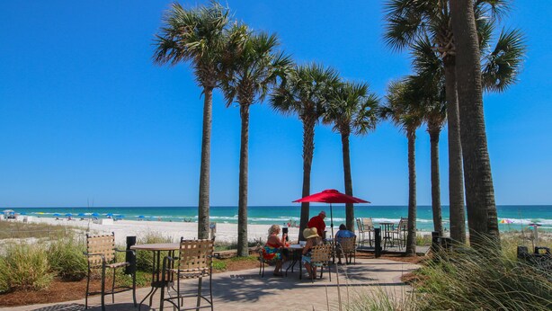 A beachside patio with tables and chairs, palm trees, and a view of the ocean. - L👀K!!! ⭐️ TOP RATED CONDO ⭐️ 3 Pools ⭐️ Breathtaking Ocean Views !!! (Panama City Beach)