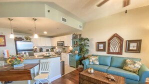A modern kitchen with white cabinets, a central island, and a dining area with a wooden table and white chairs. A teal sofa and a wicker coffee table are in the living area. - L👀K!!! ⭐️ TOP RATED CONDO ⭐️ 3 Pools ⭐️ Breathtaking Ocean Views !!! (Panama City Beach)