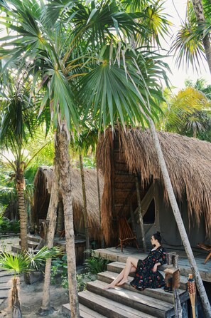 A woman sitting on wooden steps in a tropical setting with thatched-roof huts and palm trees. - Nativus Glamping & Cenote Tulum (Tulum)