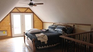 Quarto com teto inclinado, cama com roupa de cama estampada, ventilador de teto e piso de madeira.