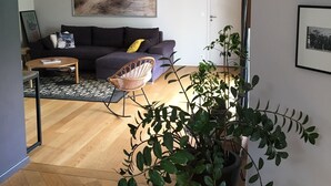 Een moderne woonkamer met een donkergroene bank, een ronde salontafel en een groot schilderij aan de muur. In de voorgrond staat een potplant en er is een rieten stoel.