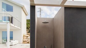 Un espace de douche extérieur moderne, doté d’un sol carrelé en pierre, d’une pomme de douche en métal et d’un tuyau d’arrosage.