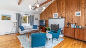 Un salon avec un plafond en bois, une cheminée en briques blanches, un canapé, un téléviseur et un meuble en bois orné d’objets décoratifs.