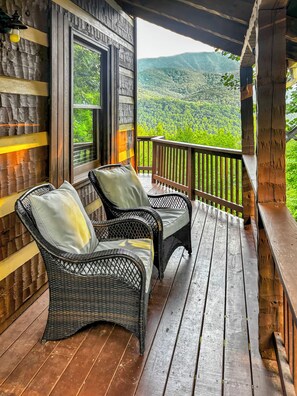 Una terraza de madera con sillas de mimbre y vista a una cadena montañosa boscosa.
