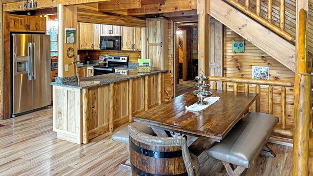 A rustic cabin interior with a wooden staircase, a kitchen with stainless steel appliances, and a dining area with a wooden table and bench.