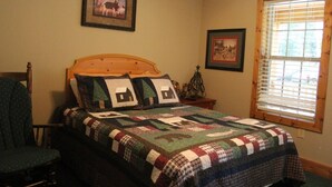 Um quarto com cabeceira de madeira, colcha estampada com motivos de casas, uma cadeira e duas molduras com quadros na parede.