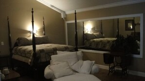 Un lit à baldaquin, un coin salon avec un pouf blanc et un grand miroir qui reflète la pièce.