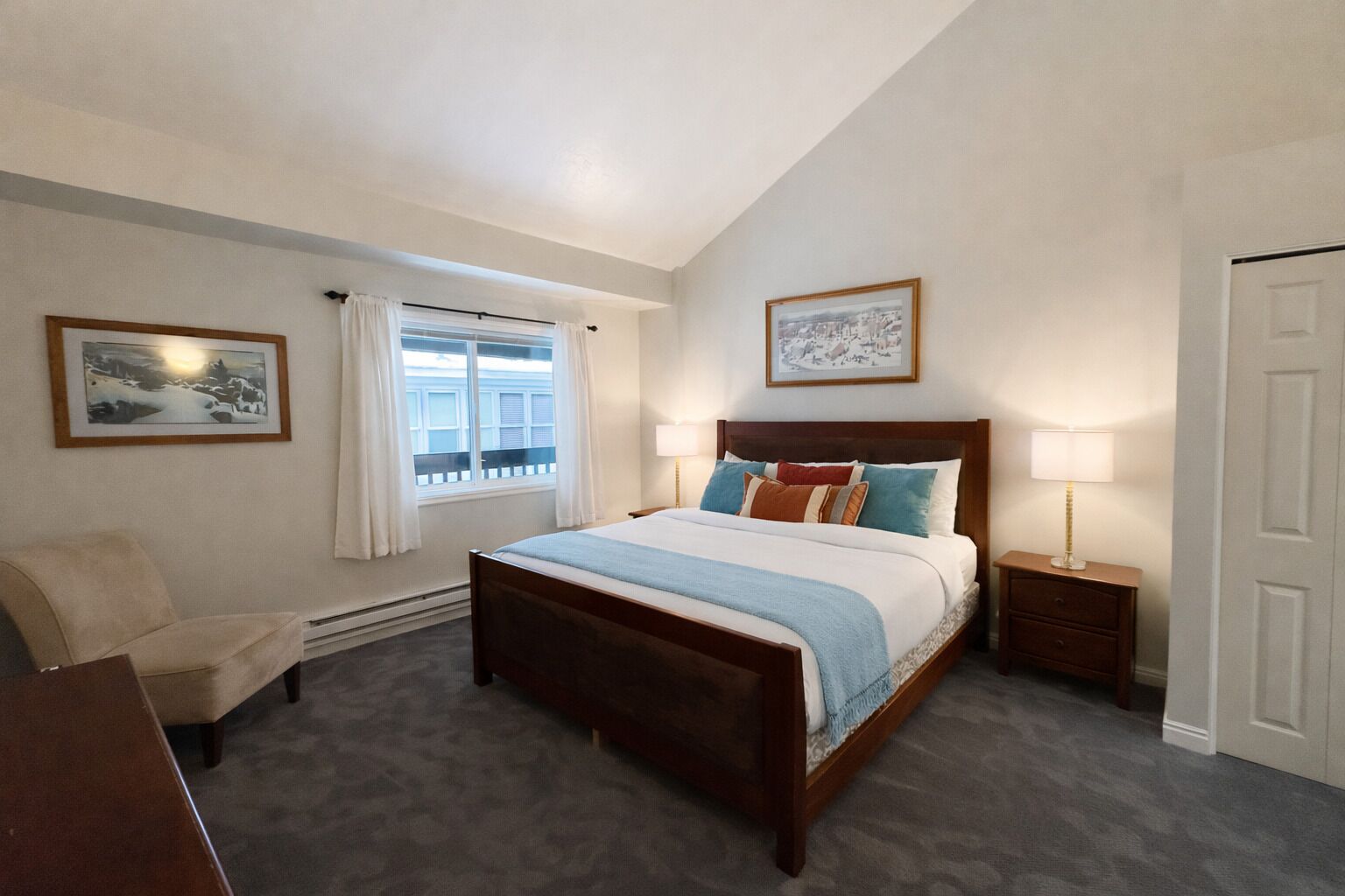 Une chambre avec un grand lit, une chaise, une table de chevet avec une lampe, une fenêtre avec des rideaux et deux tableaux encadrés accrochés au mur.