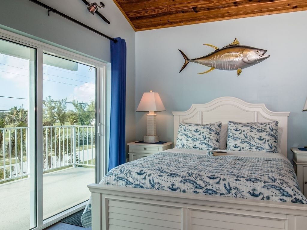 A bedroom with a large bed, a nightstand with a lamp, a wall-mounted fish decoration, and a balcony with a view of the outdoors.