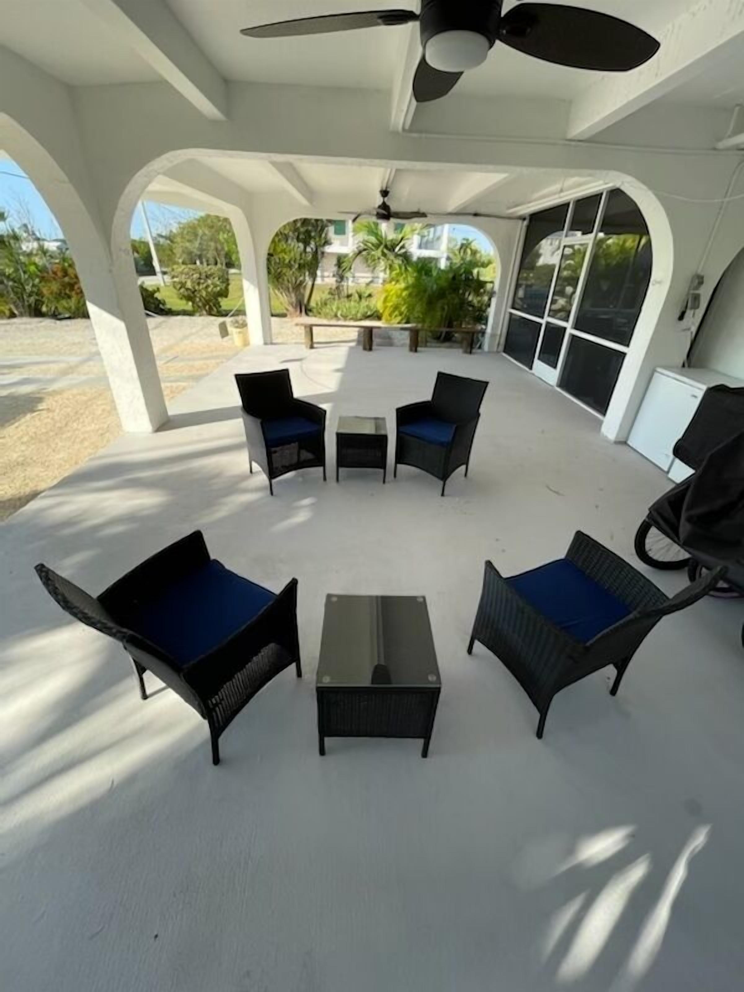 An outdoor patio with four black wicker chairs with blue cushions, a small round table, and a ceiling fan.