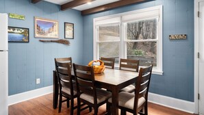 A dining area with a wooden table and chairs, framed pictures on the wall, and a window with a view of trees.