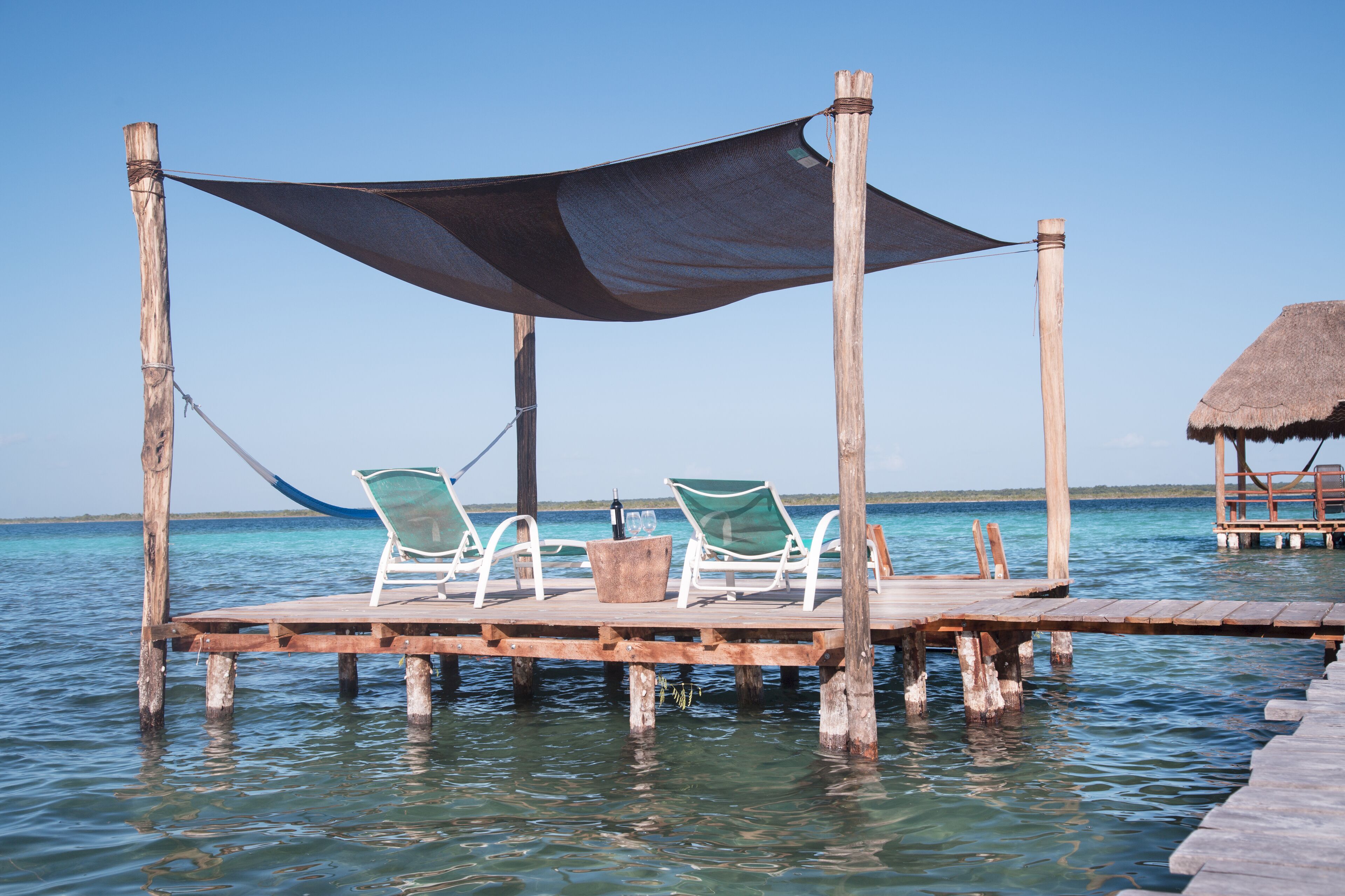 Un muelle de madera con dos sillas reclinables blancas bajo una zona sombreada, que se adentra en aguas azul cristalino.