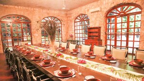 Una larga mesa de comedor dispuesta con platos, cuencos y vasos, flanqueada por sillas, en una habitación con ventanas de arco y una estantería de madera.