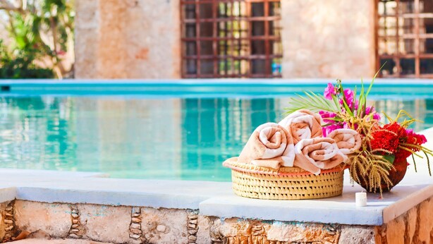 Escena junto a la piscina con una cesta tejida que contiene toallas y un jarrón con flores.