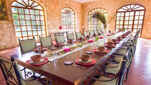Una larga mesa de comedor dispuesta para una comida en una habitación con paredes de piedra y ventanas arqueadas amplias.