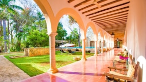 A covered outdoor area with a tiled floor, wooden benches, and a view of a garden with trees and a pool. - HACIENDA Blanca Flor CAMINO REAL (Hecelchakan)