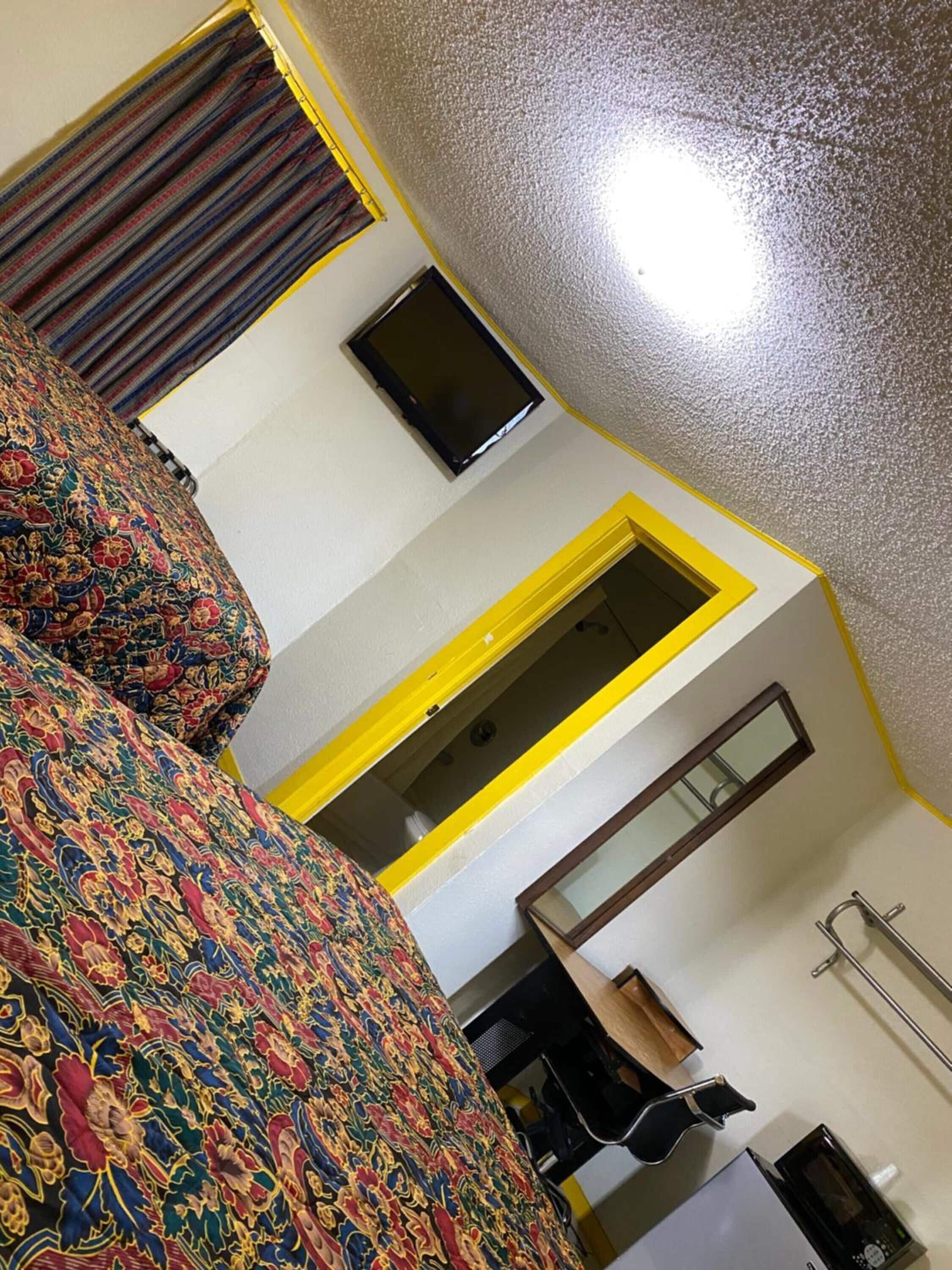 A room with a bed, a television mounted on the wall, a small desk with a chair, and a microwave.