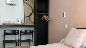 Habitación de hotel con cama, dos sillas, una mesa con toallas, un estante con perchas, un cuadro y un teléfono en la pared.