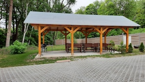 Un pavillon en bois doté de bancs, entouré de verdure et bordé d’une zone pavée.