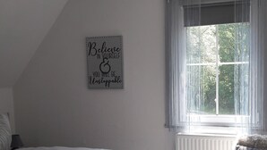 Une chambre à coucher avec un lit, une chaise, une fenêtre avec des rideaux et une citation inspirante sur le mur.