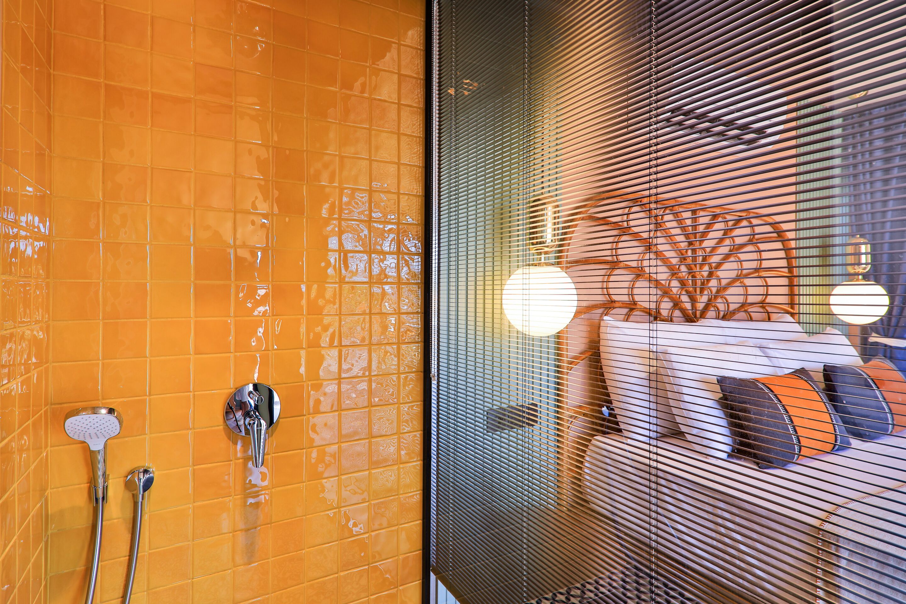 A bathroom with orange tiled walls, a shower with two handheld showerheads, and a window with blinds.