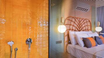A bathroom with orange tiled walls, a shower with two handheld showerheads, and a window with blinds.